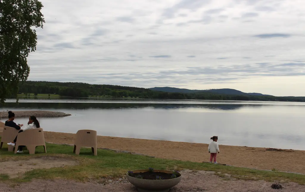 Badstranden på Långnäs, Folkets Park, Bollnäs. Gräsmatta, sandstrand, vy över sjön Varpen med skog och berg i bakgrunden.